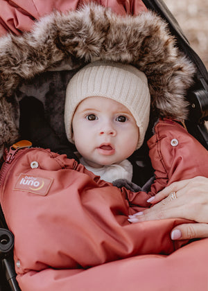 Baby girl in a rust terracotta baby luno footmuff with fur hood, wearing a white knit hat.