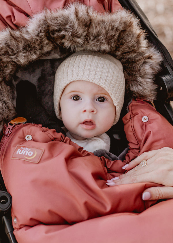 Baby girl in a rust terracotta baby luno footmuff with fur hood, wearing a white knit hat.