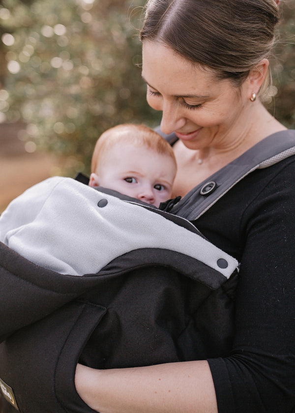 Woman carrying a baby in a black and gray baby carrier winter cover outdoors.