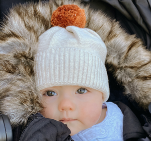 Baby in a stroller with a fur-lined hood, wearing a white knit hat with a pom-pom, and a baby-specific liner. The stroller has 'baby luno' branding.
