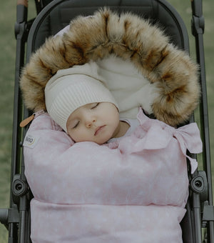 Baby sleeping in a stroller with a pink swaddle and brown fur hood.