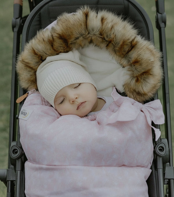 Baby sleeping in a stroller with a pink swaddle and brown fur hood.